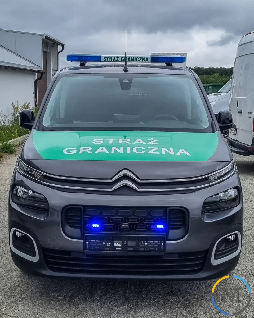 Szary radiowóz marki Citroën polskiej Straży Granicznej z zieloną maską i włączonymi niebieskimi światłami uprzywilejowania.