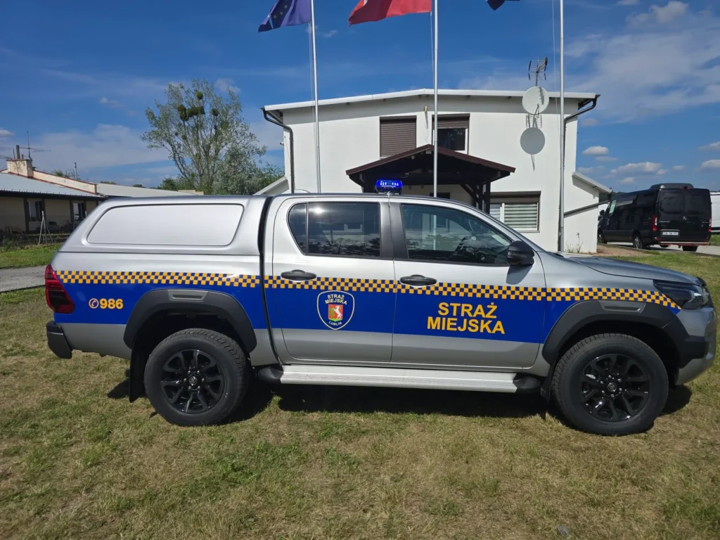 Srebrny radiowóz pickup Toyota Hilux Straży Miejskiej w Lublinie zaparkowany na trawie przed budynkiem