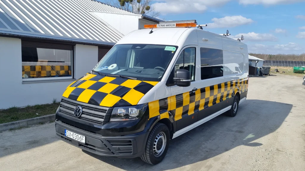 Biały Volkswagen Crafter z żółto-czarną szachownicą ostrzegawczą oraz belką świetlną z napisem Utrzymanie Nawierzchni zaparkowany przy budynku.